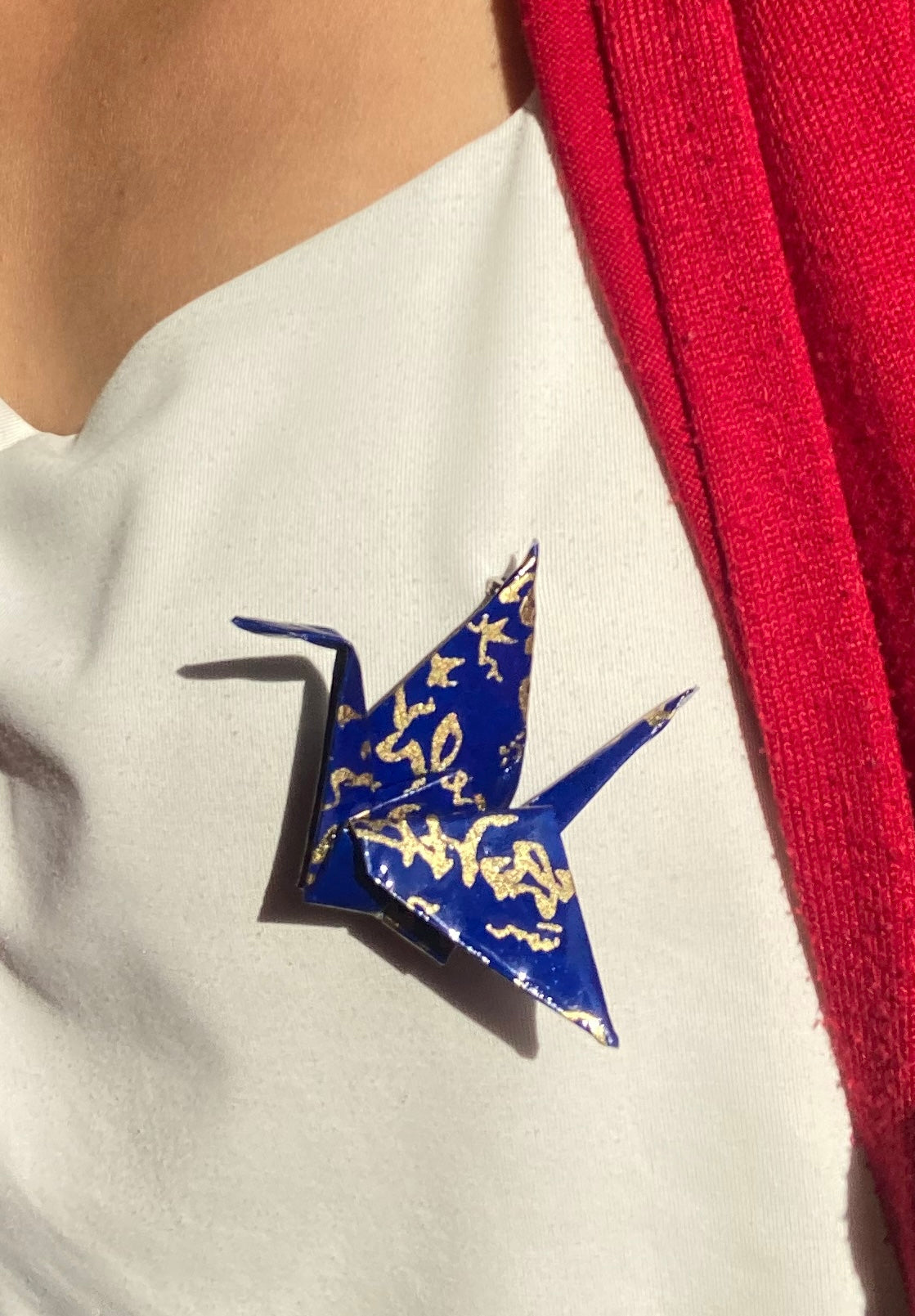 A hand-folded origami crane brooch in vibrant colors, displayed on a white surface with a pink flower and green leaves in the background.