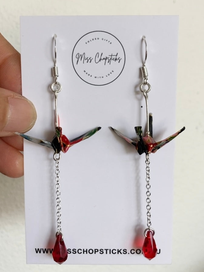 A pair of origami earrings featuring a crane design with Swarovski crystals and sterling silver components, displayed against a reflective background.