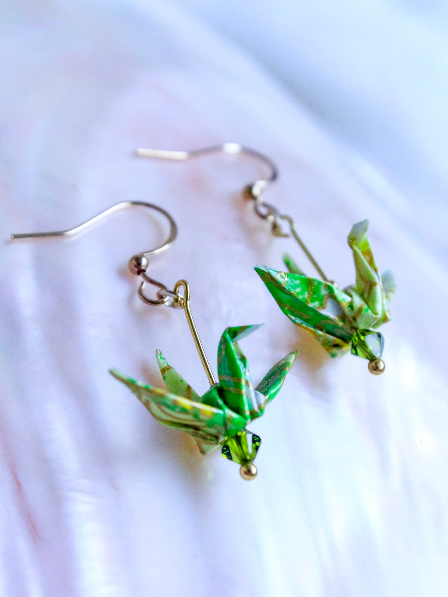 A hand-folded origami crane earrings in blue and white, perched atop a Swarovski crystal, with gold-colored hooks.