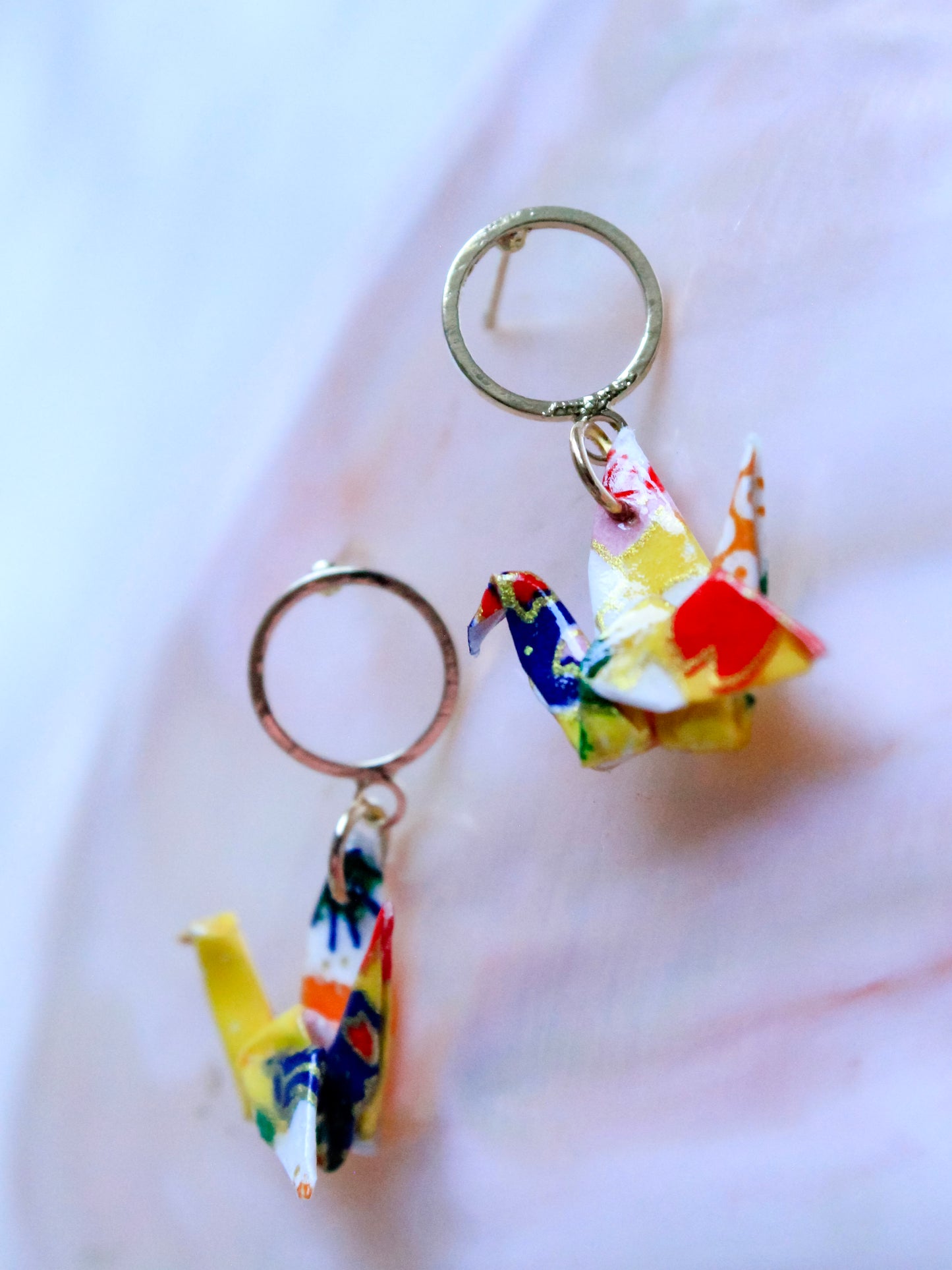 A pair of origami crane hoop earrings with multicolored patterns, displayed against a pink background.