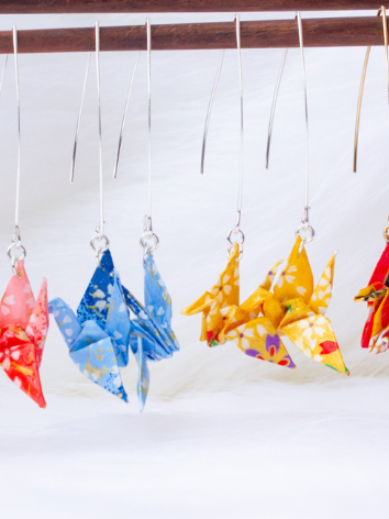 A series of origami crane earrings in various colors hang from silver hooks, displayed against a white backdrop.