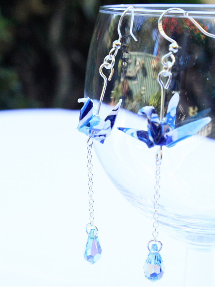 A pair of origami earrings featuring a crane design with Swarovski crystals and sterling silver components, displayed against a reflective background.