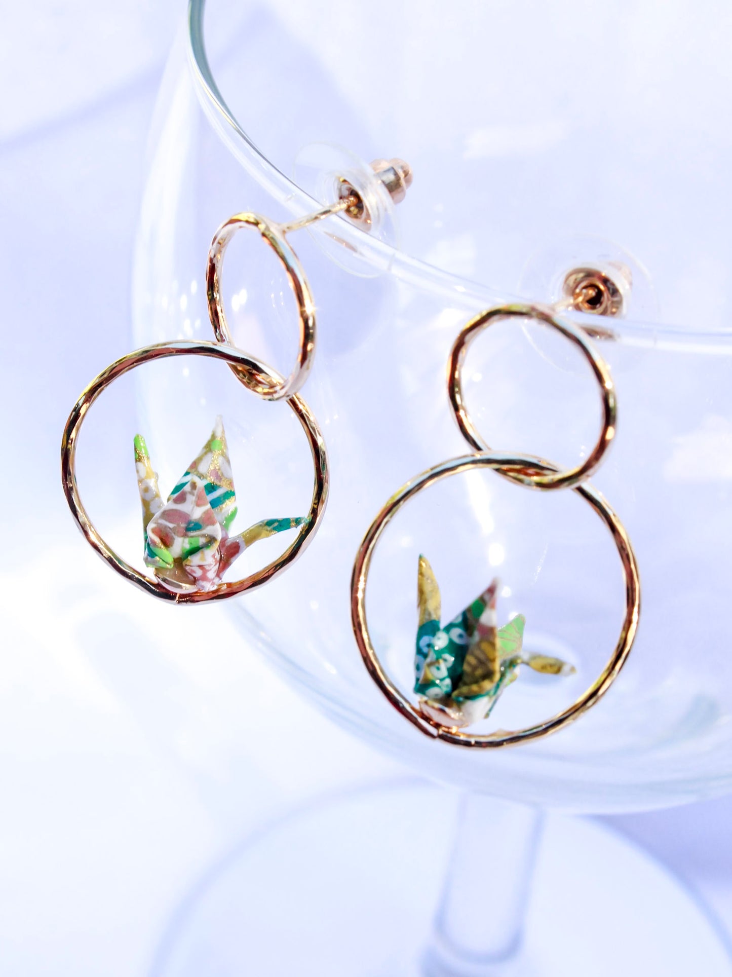 A pair of origami earrings featuring a tiny red crane inside golden double hoops, displayed against a clear glass background.