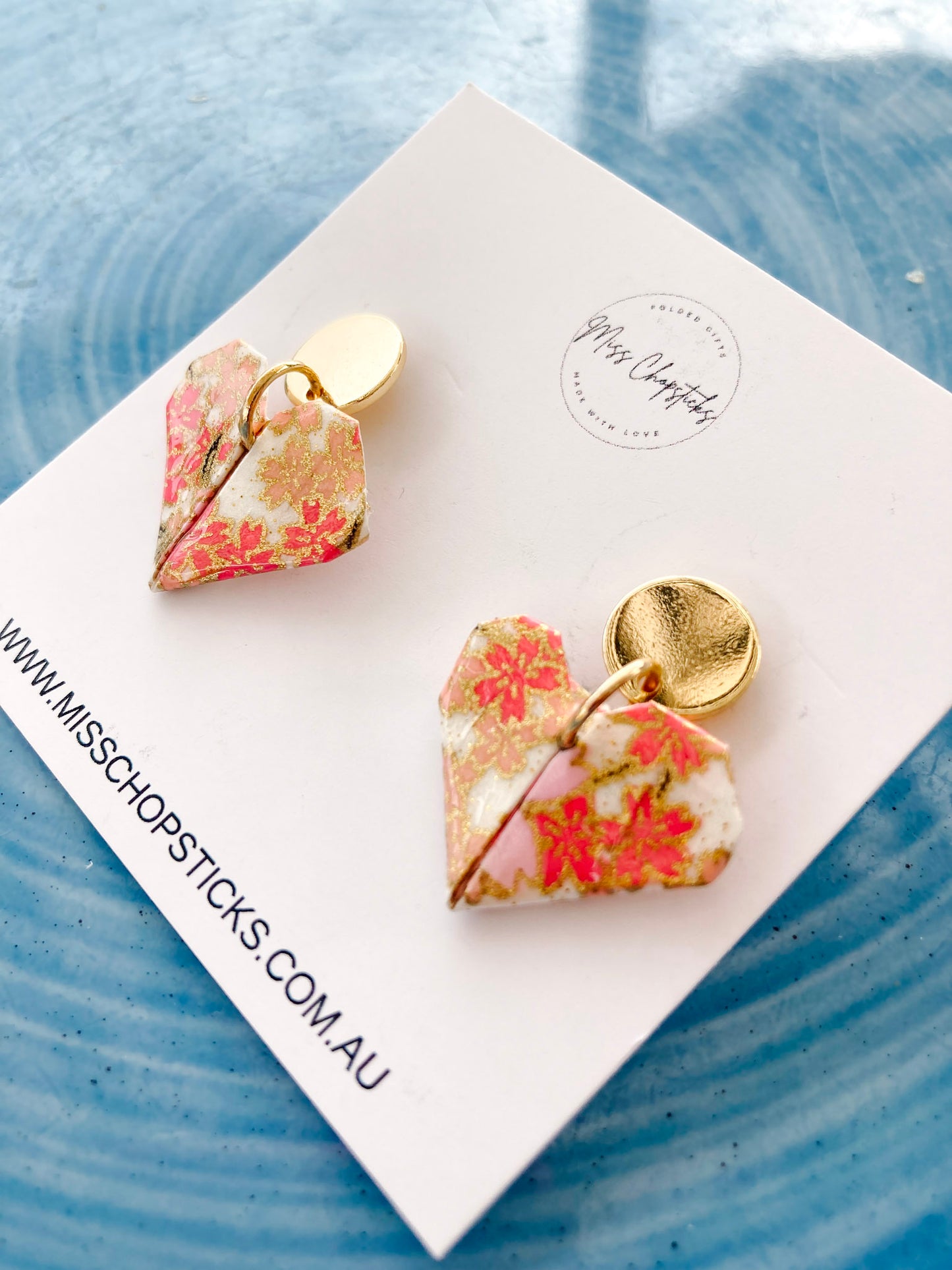 A pair of origami love heart-shaped earrings with golden studs on a paper stock card.